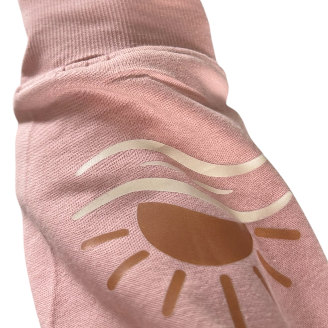 Gros plan de la manche du Crewneck Coastal rose pâle Peek Breeze avec illustration de soleil et de vagues, inspirée des paysages côtiers.